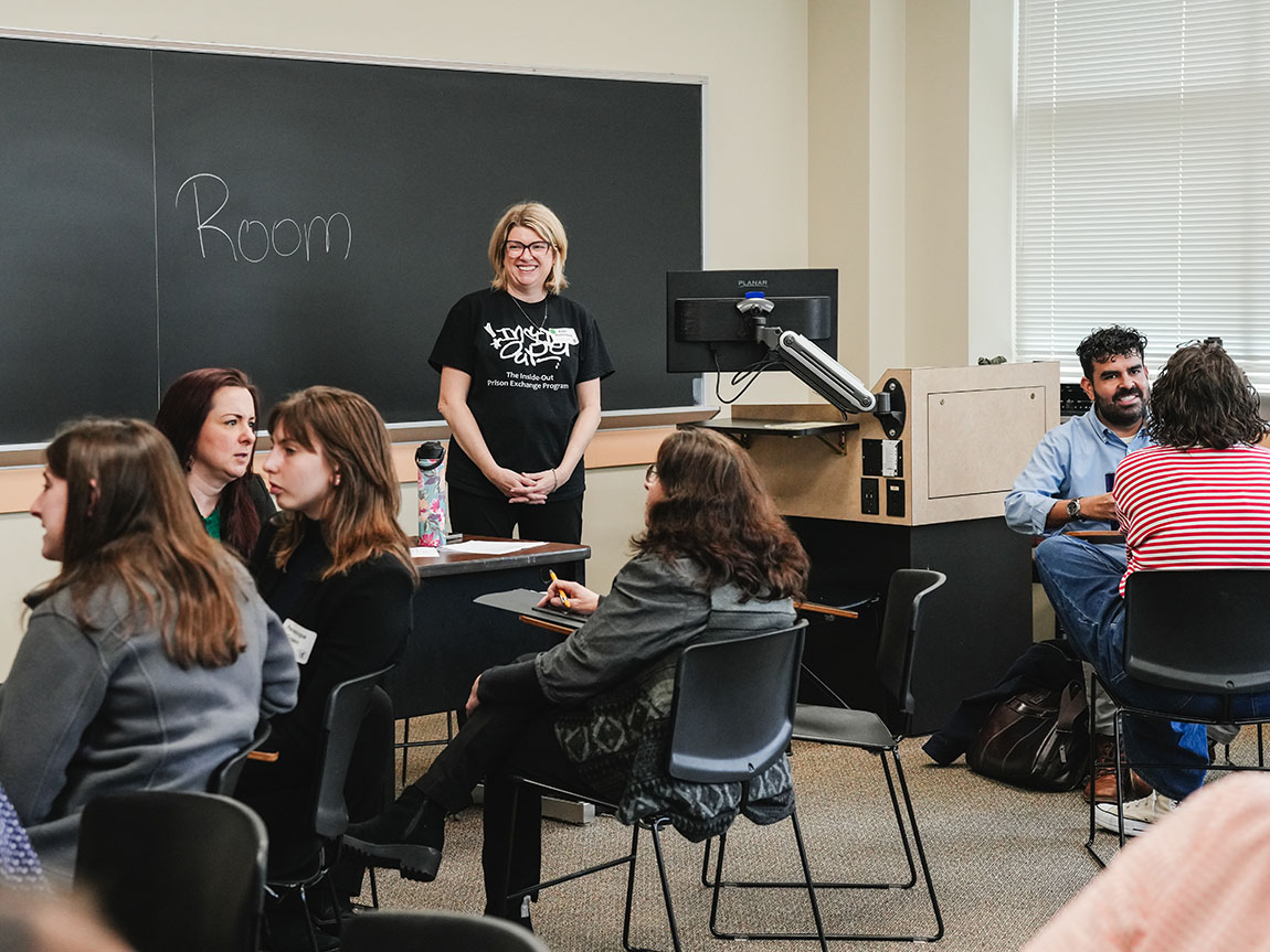 A college professor with blonde hair in a black T-shirt stands in a classroom